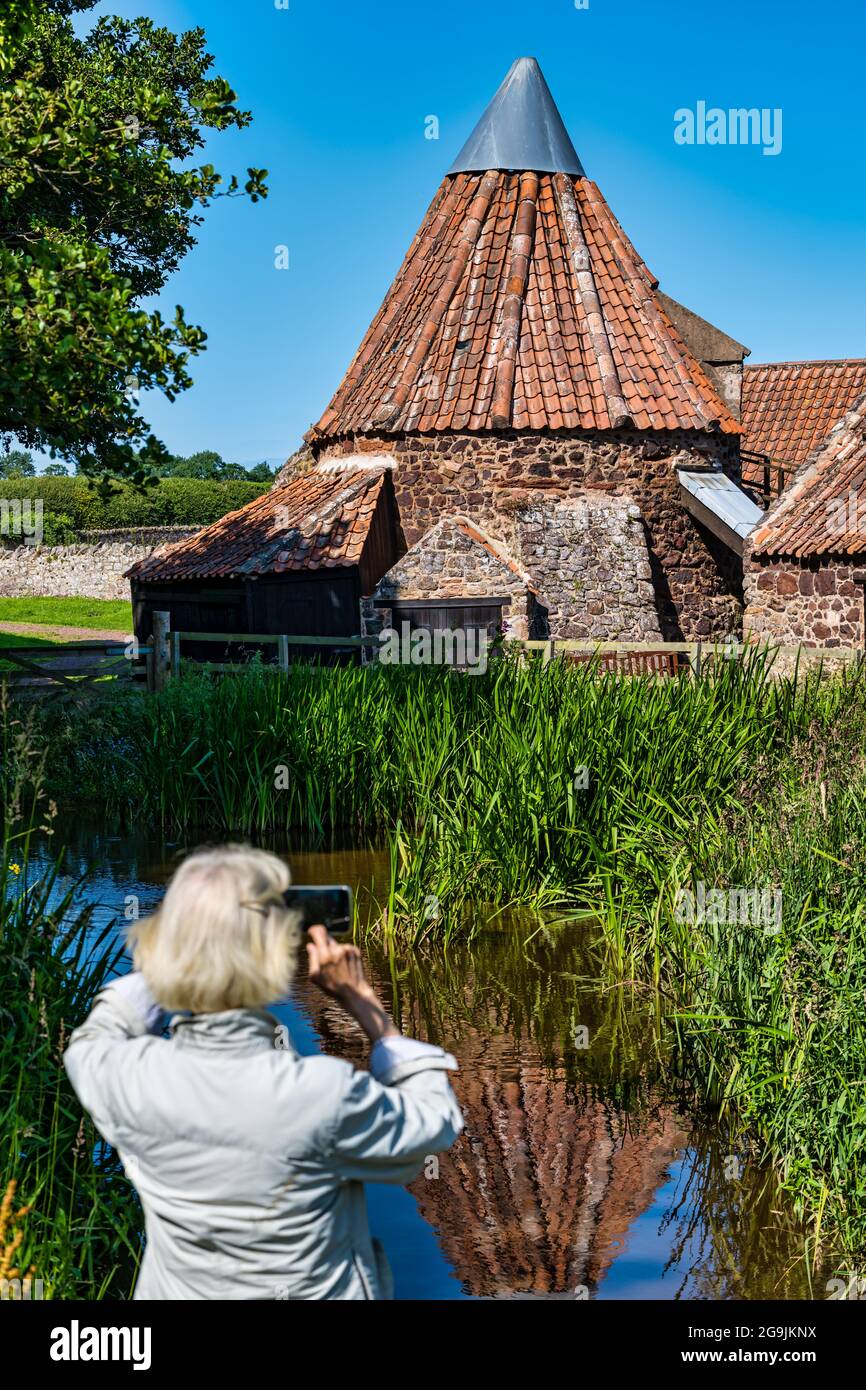 Senior woman taking photo of historic quirky old Preston Mill reflected in mill pond on sunny Summer day, East Lothian, Scotland, UK Stock Photo