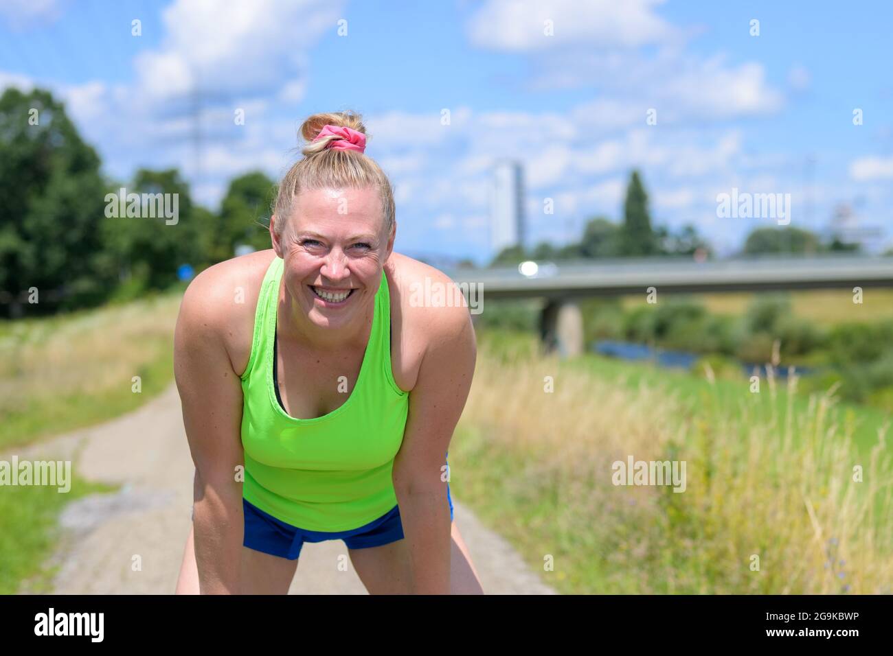 Cute happy blond woman with a lovely beaming charismatic smile bending forwards to lean on her knees as she takes a rest while jogging on a rural foot Stock Photo
