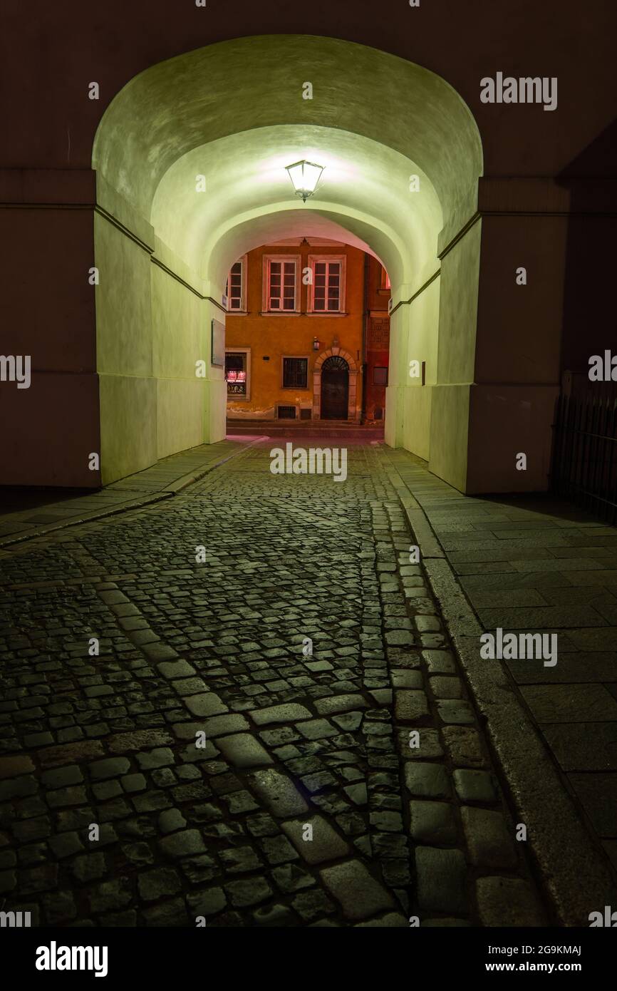 Cobblestone empty street at night with arched passage under building, illuminated with single lamp in Old Town of Warsaw, Poland. Stock Photo