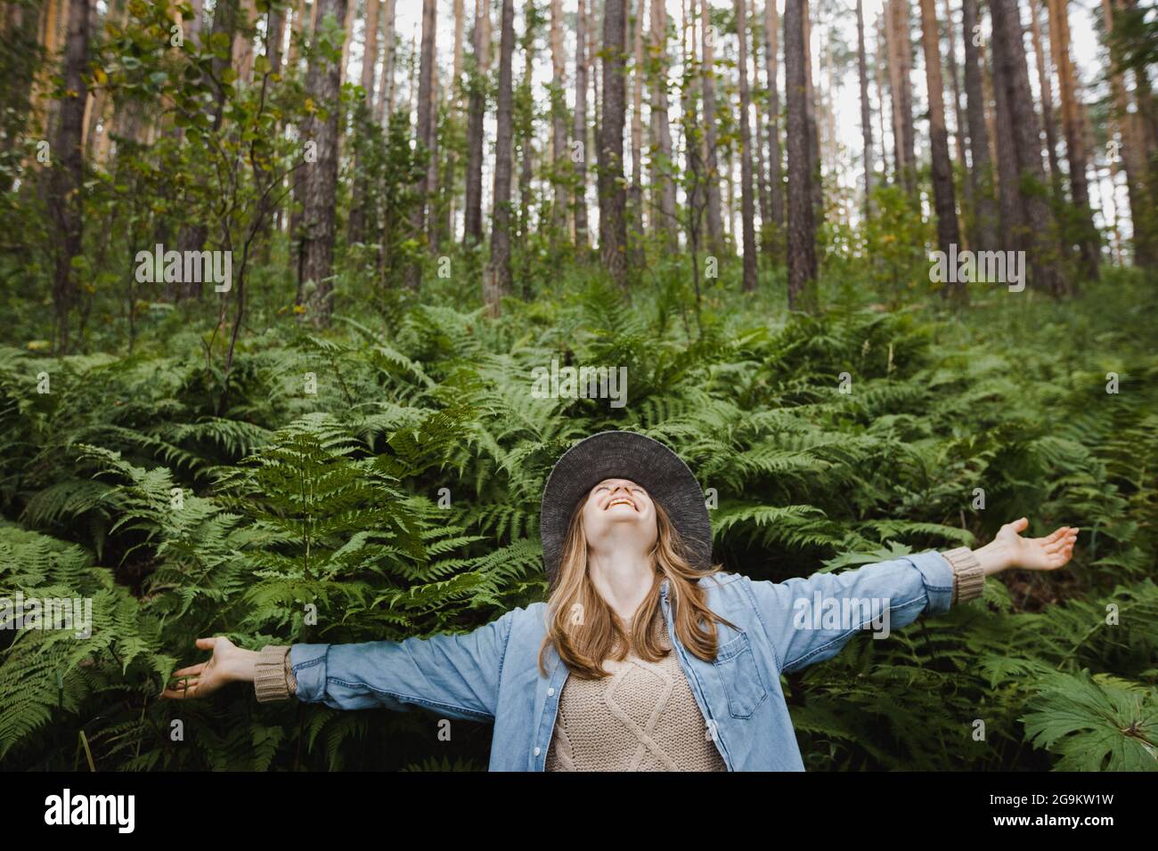 Positive female with outstretched hands in abundant fern growing in woods with tall pines Stock Photo