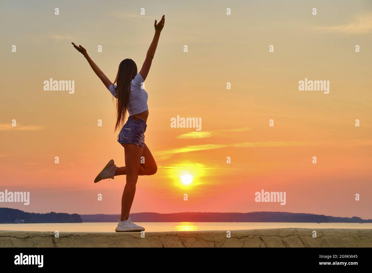 Young woman jumping and spreading hands to the sides and facing to the sunset Stock Photo
