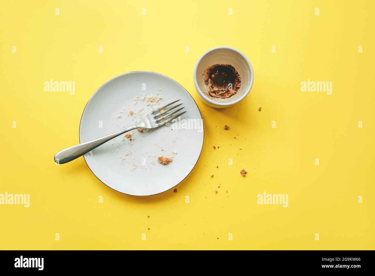 Empty plate with fork and crumbs after eating and cup with coffee grounds on yellow background. Concept of the end of the party or celebration. Stock Photo