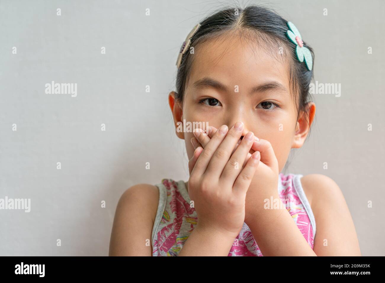 Close-up portrait of Asian little child girl with black long hair tied, covering her mouth with two hands. Stock Photo
