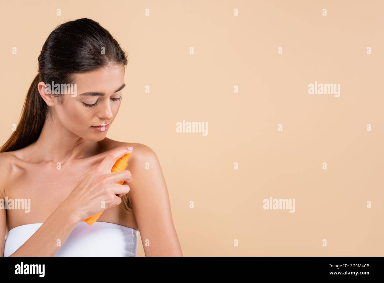 brunette woman spraying sunblock on shoulder isolated on beige Stock Photo