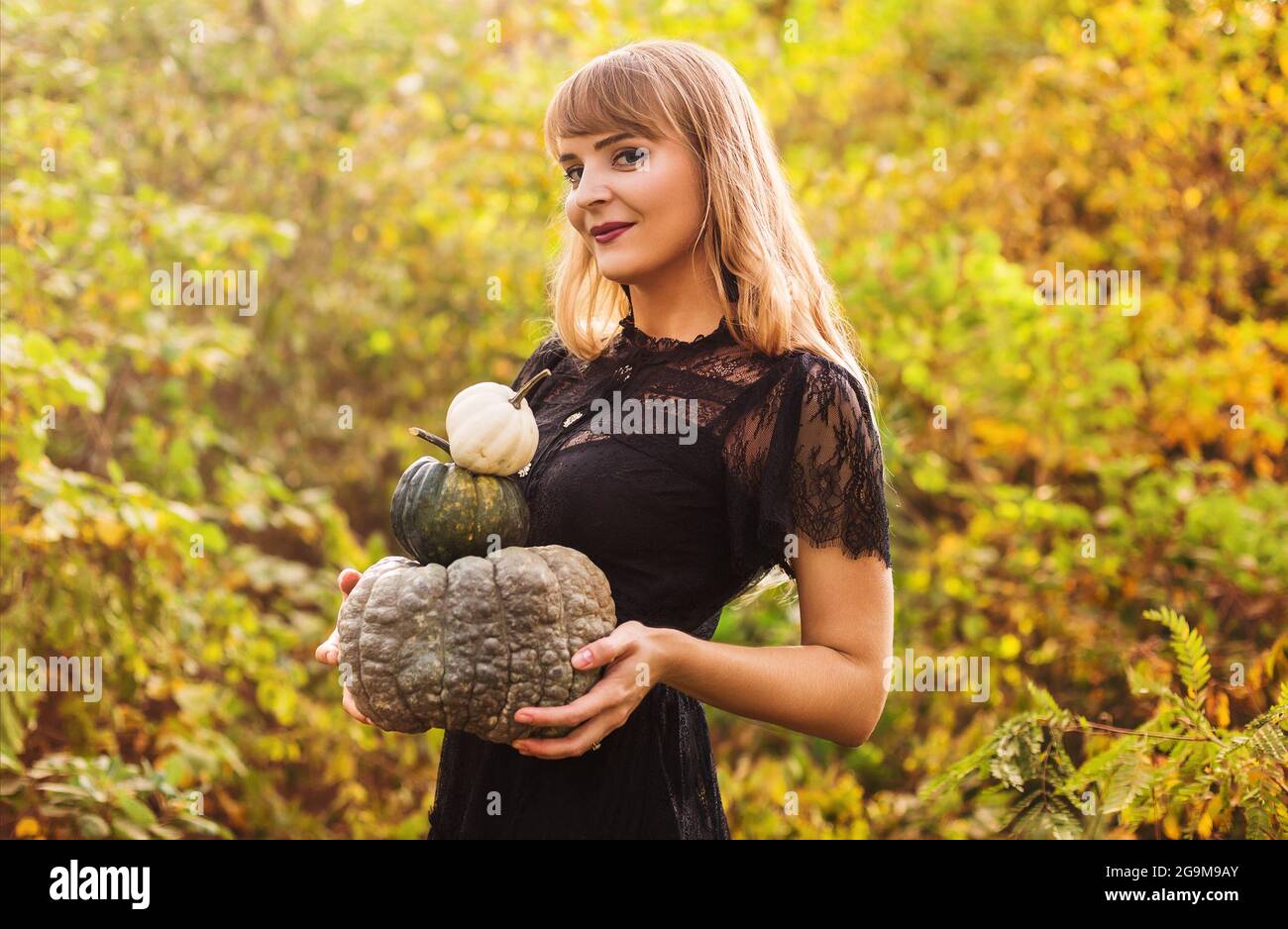 Portrait of beautiful young blond woman with long straight hair and dark vamp makeup looking to the camera, wearing black guipure dress, holding pumpk Stock Photo