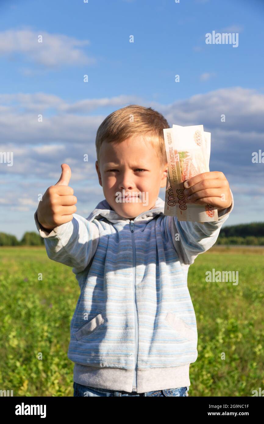 A satisfied preschooler child in a sweater holds paper rubles in nature in a field against the background of a blue sky with clouds. Close-up. Portrai Stock Photo