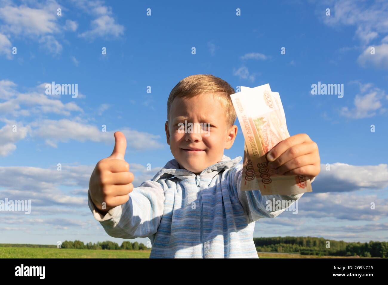 A satisfied preschooler child in a sweater holds paper rubles in nature in a field against the background of a blue sky with clouds. Close-up. Portrai Stock Photo