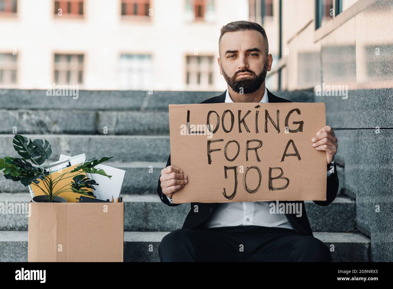 Looking for a job. Upset mature man sitting on stairs with box of personal stuff and poster, got fired due to crisis Stock Photo