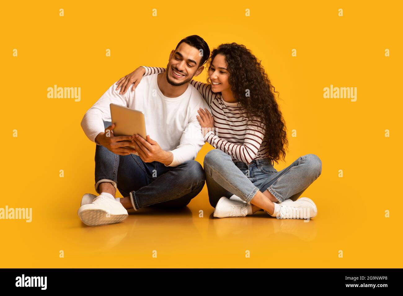 Happy Young Middle-Eastern Couple Using Digital Tablet While Sitting Over Yellow Background Stock Photo