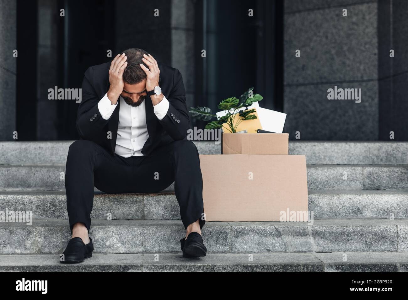 Upset mature businessman sitting with empty poster cardboard sign and box of personal stuff on stairs of office center Stock Photo