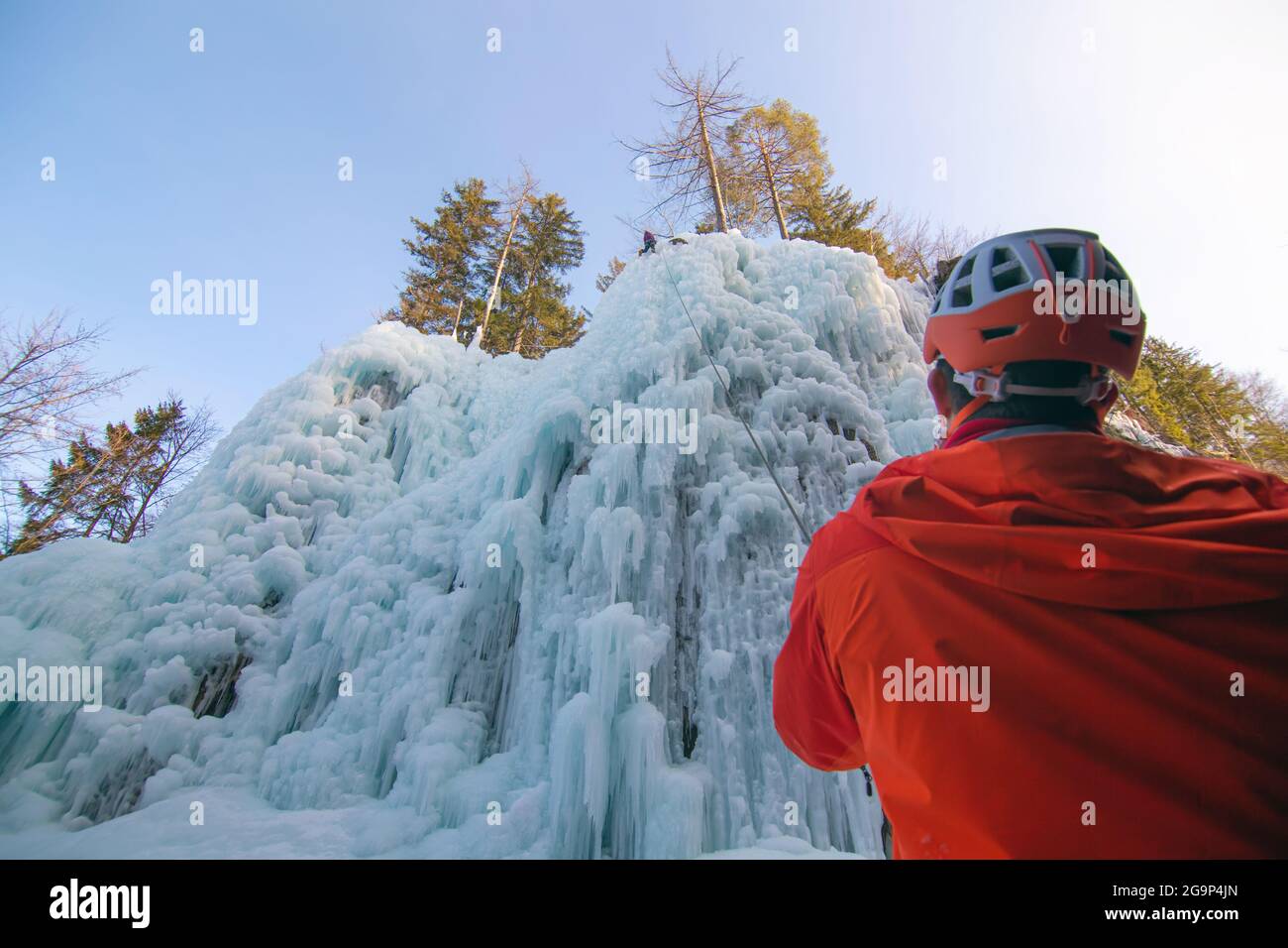 Man standing and controlling a safety top rope while female with ice climbing equipment, climbing on a frozen waterfall Stock Photo