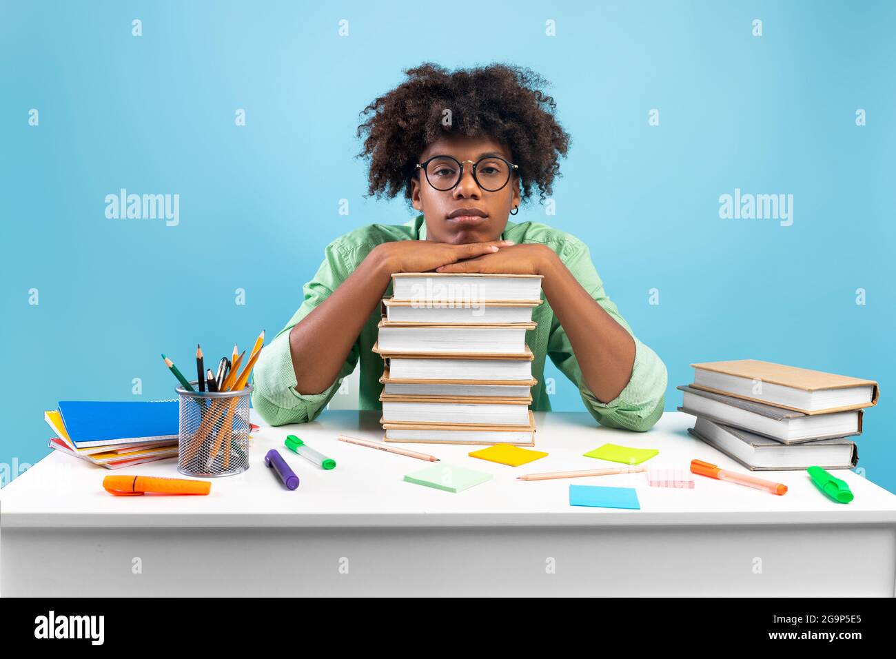 Lack of motivation. Bored african american teen guy sitting at table and leaning on stack of books, blue background Stock Photo