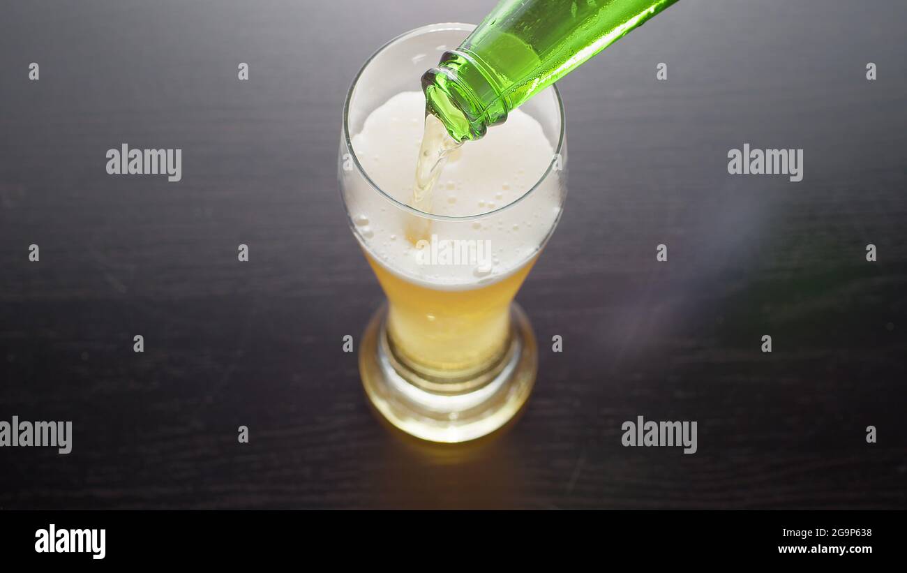 Pouring beer into glass from bottle, view from above. Half pint of frothy fresh lager beer on table. Top view. Stock Photo