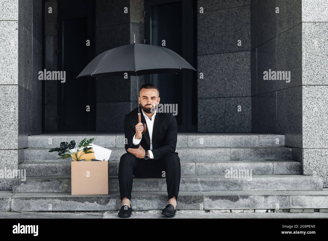 Unemployment concept. Jobless mature businessman sitting under umbrella with empty poster and box of belongings Stock Photo