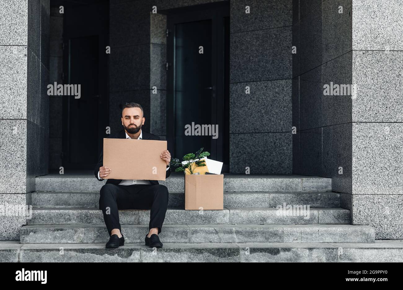 Unemployment concept. Depressed mature businessman sitting with empty cardboard sign and box of personal belongings Stock Photo