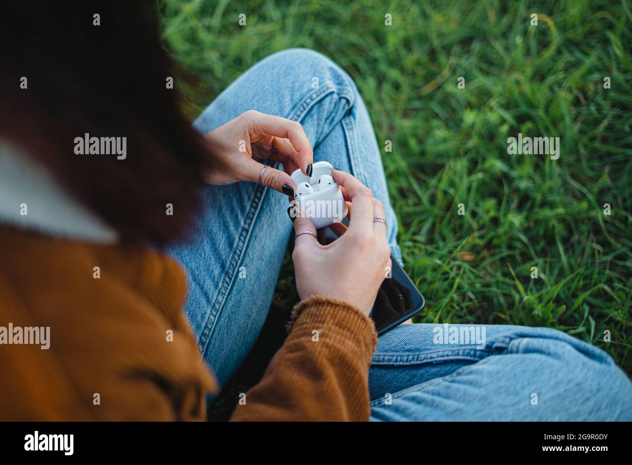 A woman opening the charging case of white wireless ear-buds while sitting on the grass (Horizontal) Stock Photo