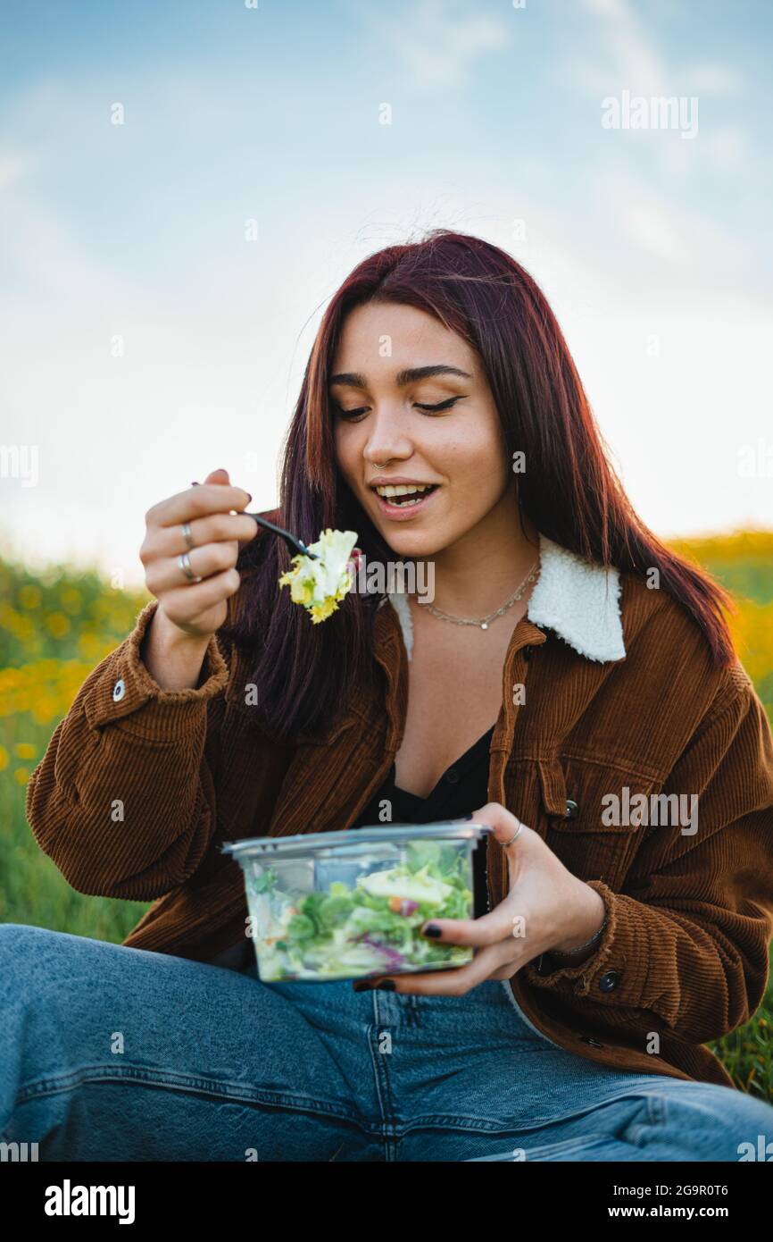 Teenager girl eating a salad while sitting on grass at a hill. She is enjoying the calm. Medium shot. Stock Photo
