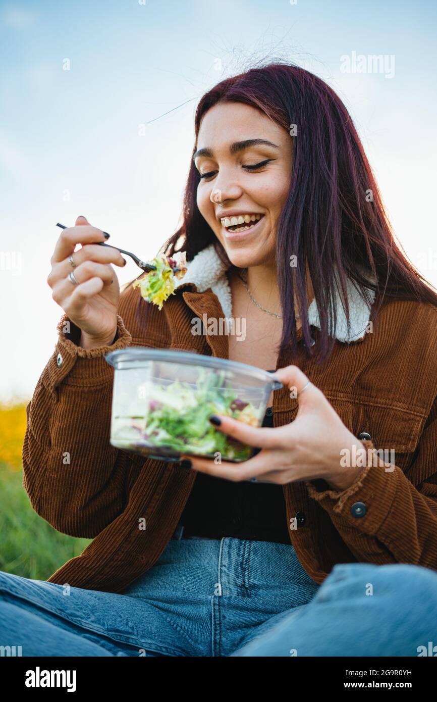 Laughing teenager girl enjoying eating a salad. She is in the field during sunset. Stock Photo