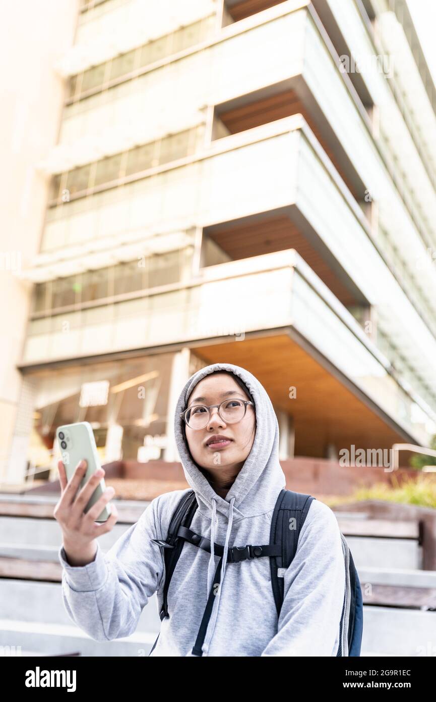 Young Asian person holding cell phone looking away from camera in city Stock Photo