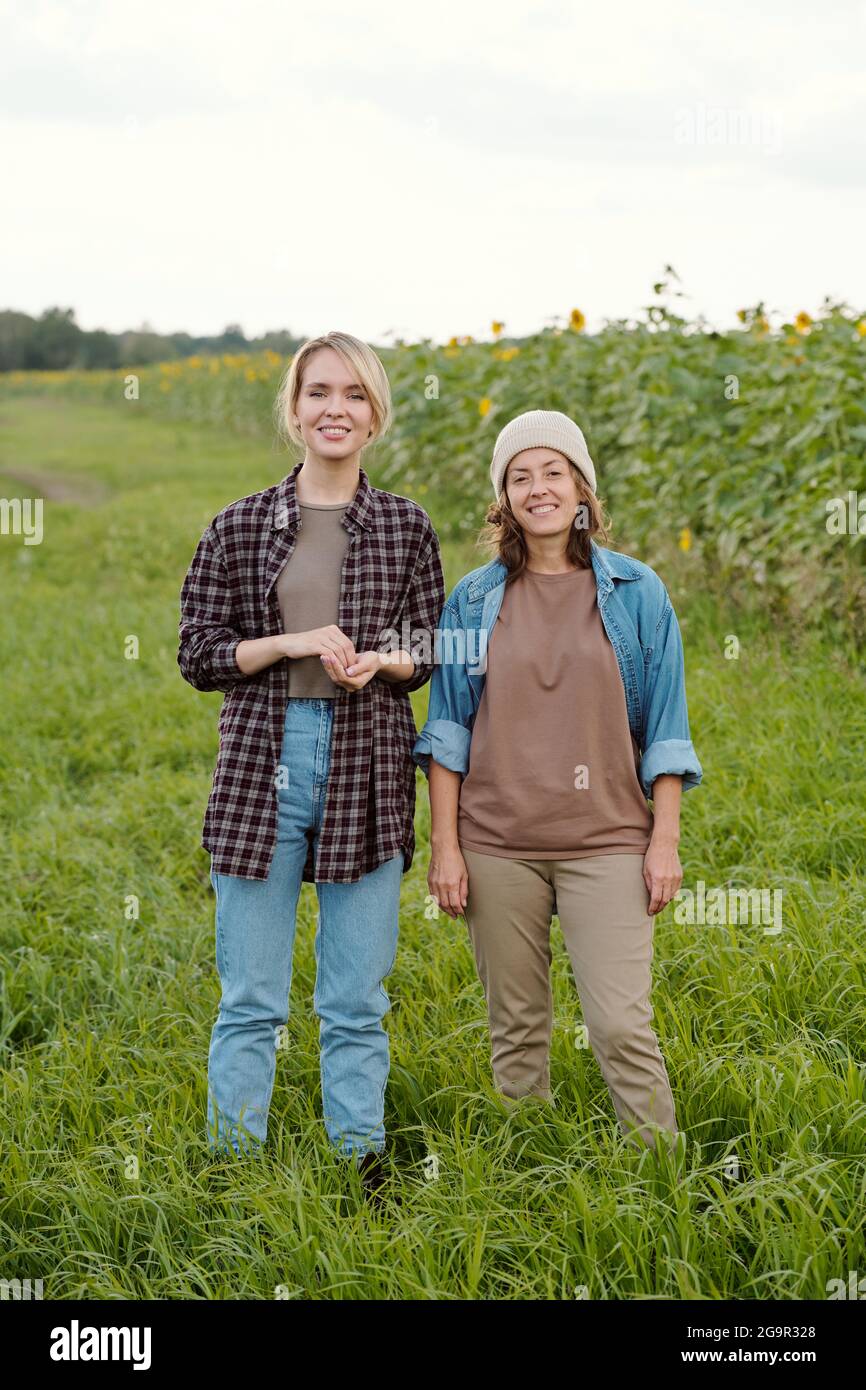 Two cheerful female farmers in workwear looking at you with smiles while standing in front of camera against green field and country road Stock Photo