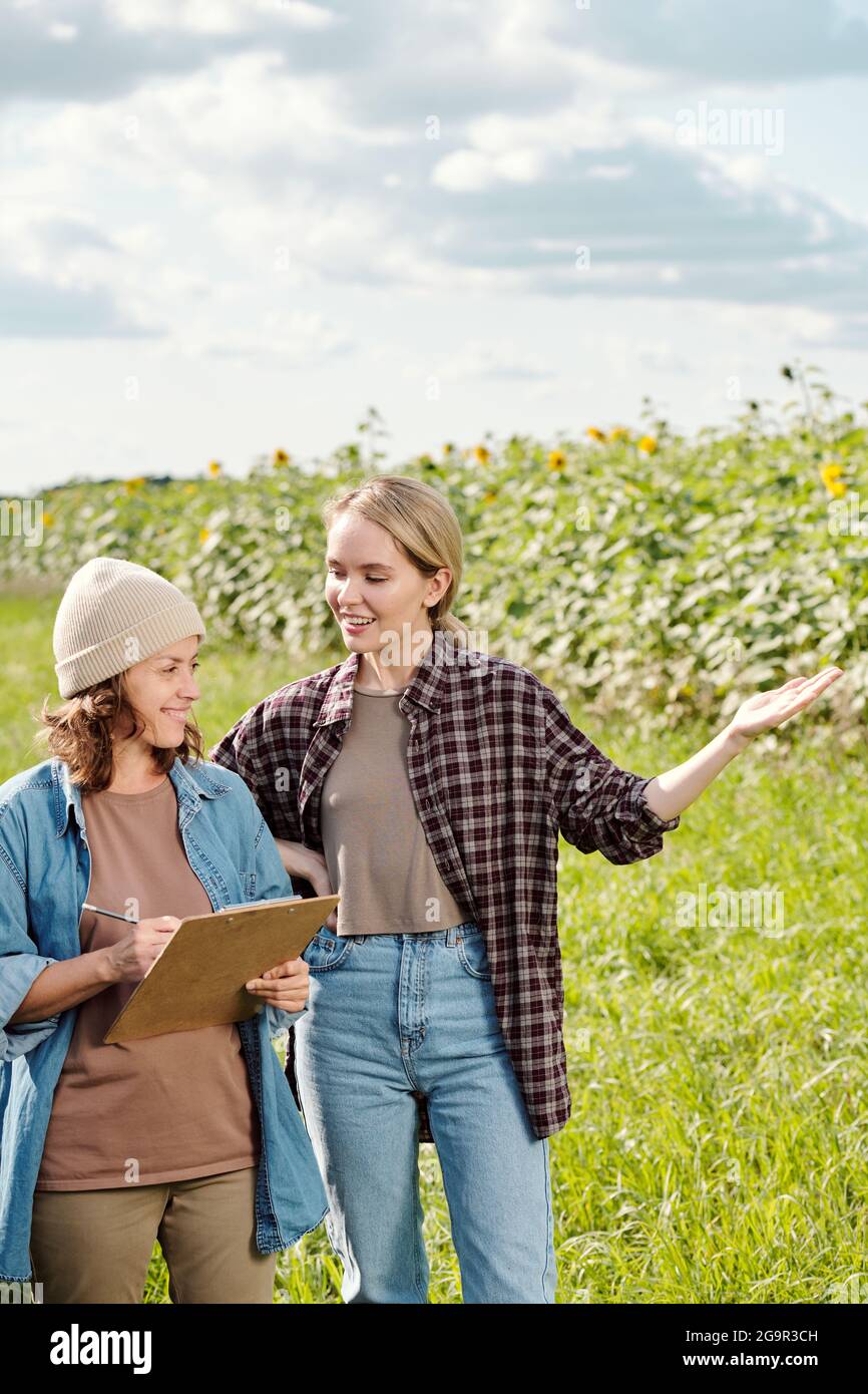 Two cheerful female farmers in workwear looking at sunflower field in front of camera while one of them making notes during discussion Stock Photo