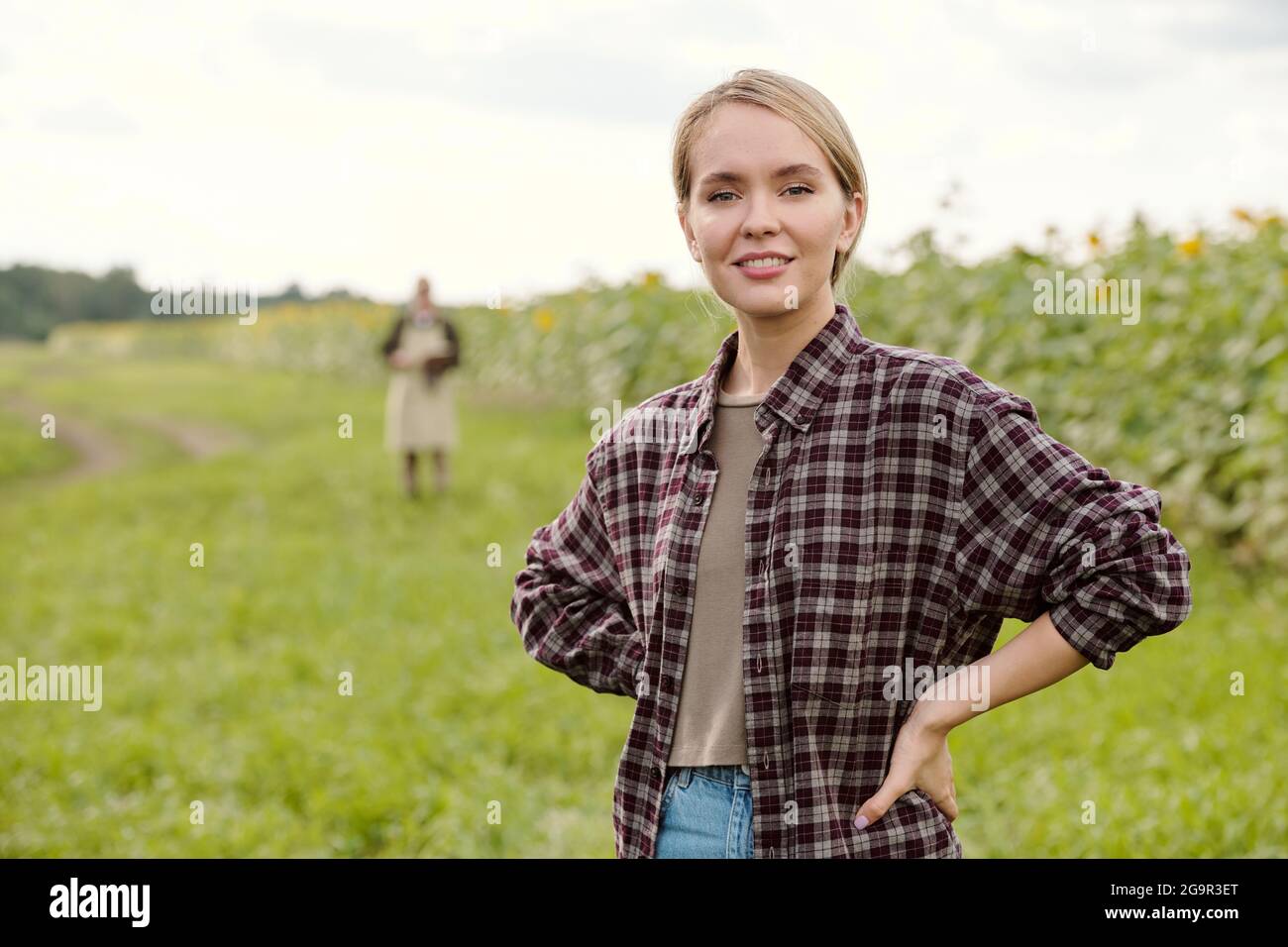Young blond smiling female farmer in workwear looking at you while standing in front of camera and using digital tablet against green field Stock Photo