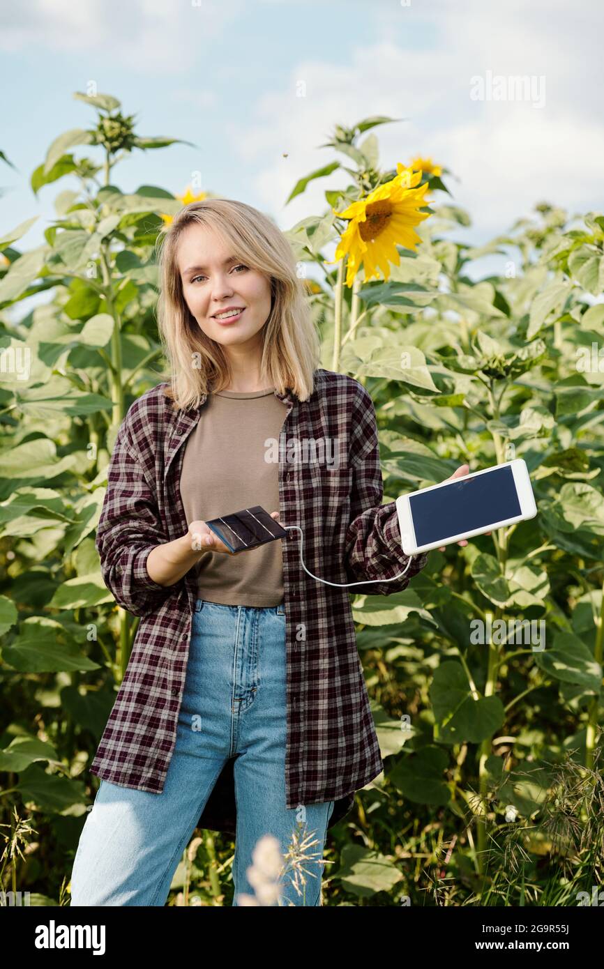 Young blond smiling female farmer in workwear looking at you while standing in front of camera and using digital tablet against green field Stock Photo