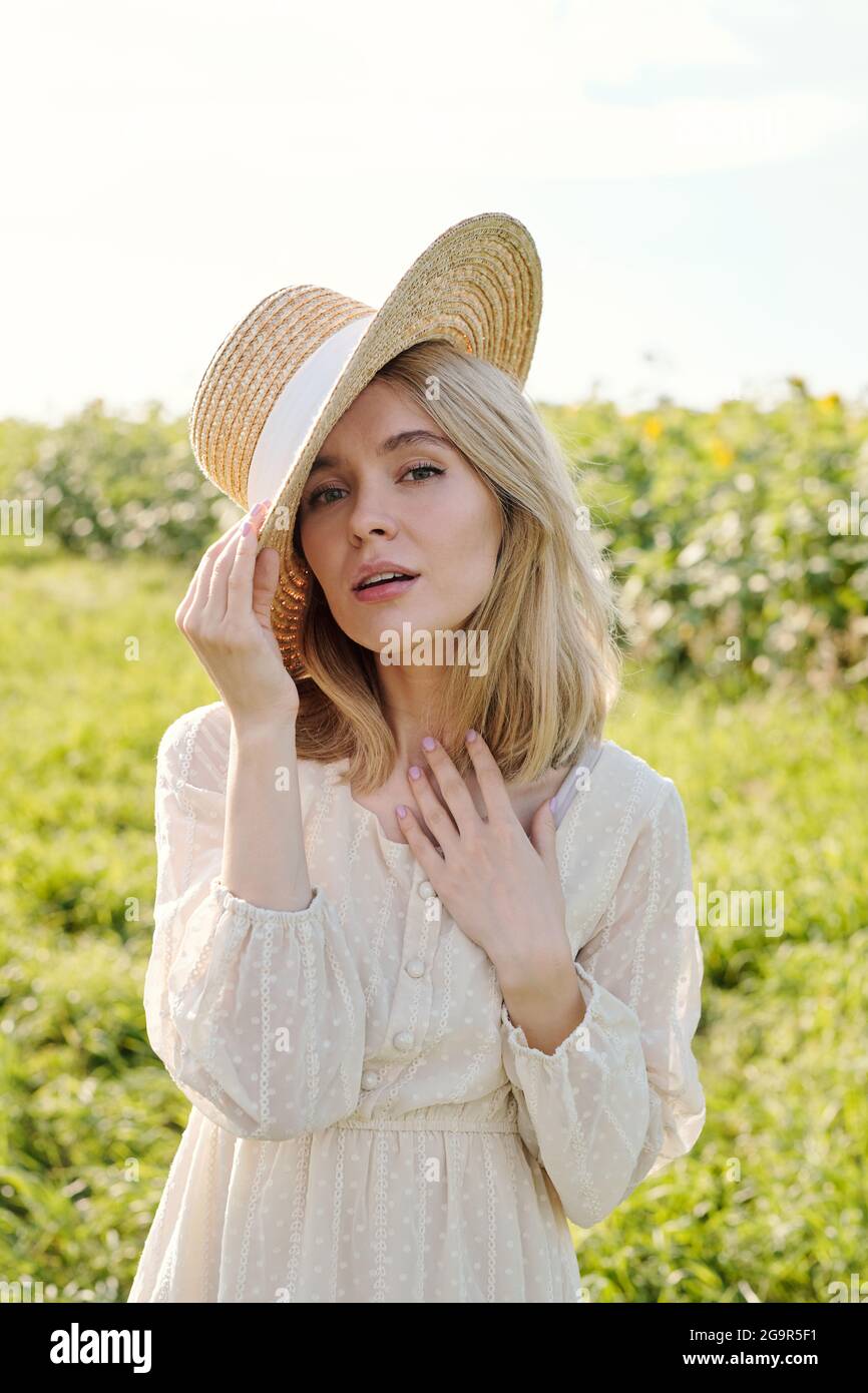 Gorgeous young blond woman in straw hat and white country style dress standing in front of camera against green grass and sunflower field Stock Photo
