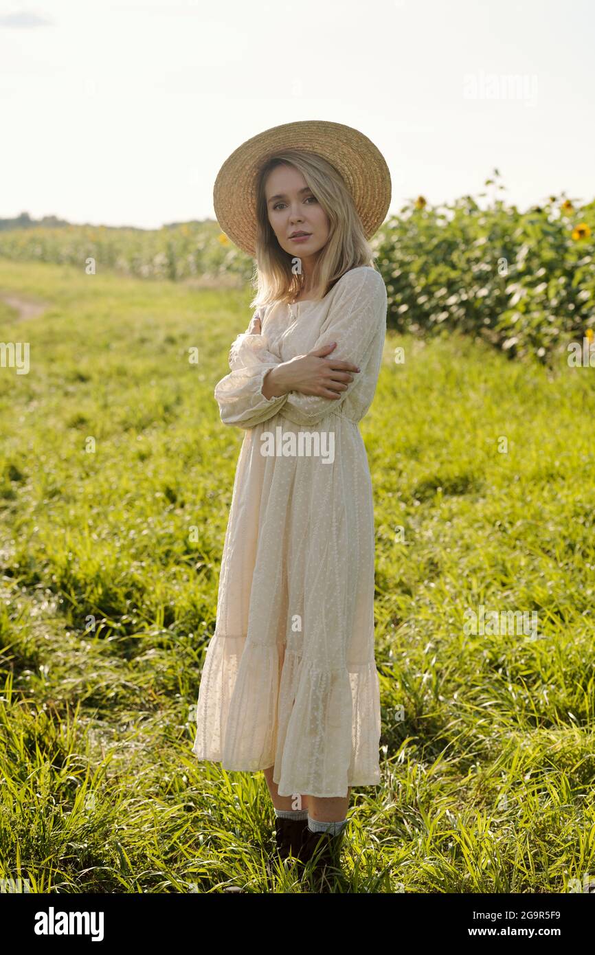 Gorgeous young blond woman in straw hat and white romantic dress standing in front of camera against sunflower field on sunny day Stock Photo