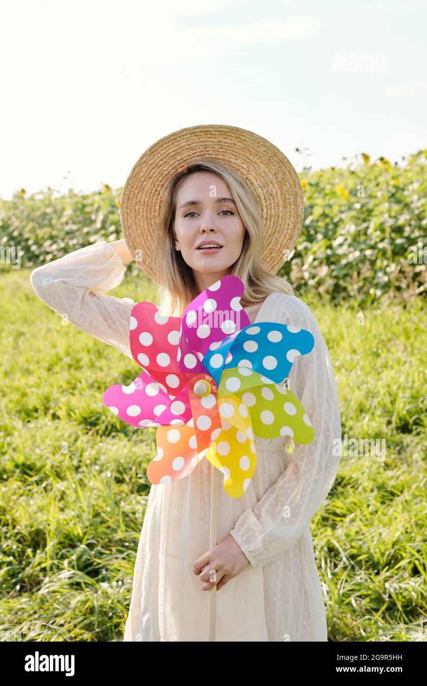 Gorgeous young blond woman in hat and country style dress holding large polkadot whirligig while standing against green grass and sunflower field Stock Photo