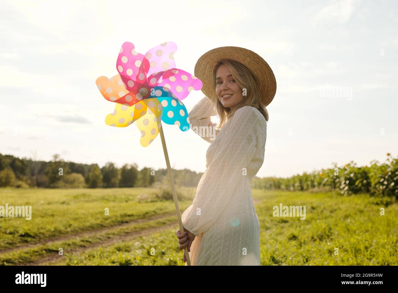 Back view of young elegant woman in hat and country style dress holding large polkadot whirligig while standing in front of green grass and road Stock Photo