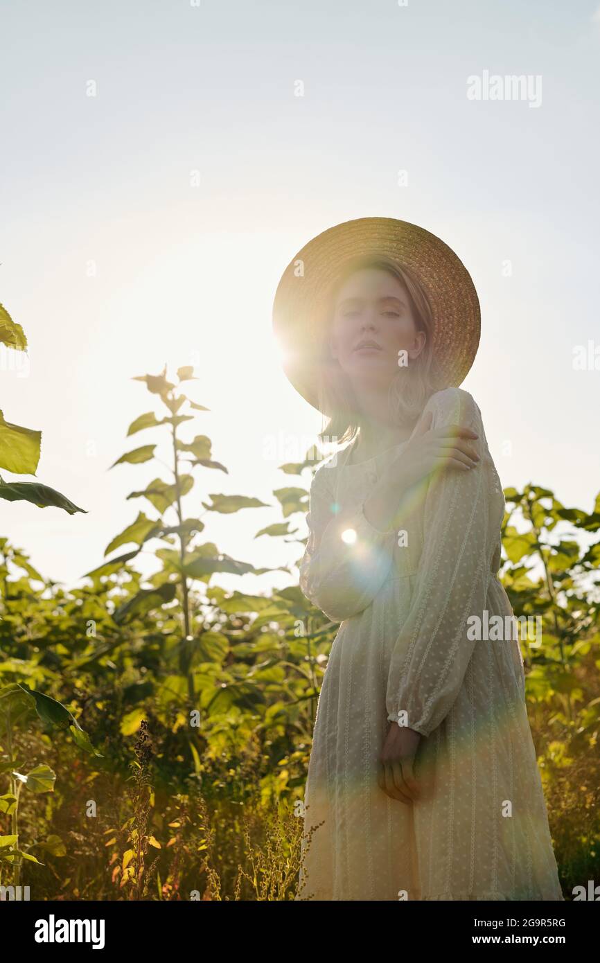 Beautiful young blond woman in straw hat and white country style dress standing in front of camera against sunflower field over cloudy sky Stock Photo