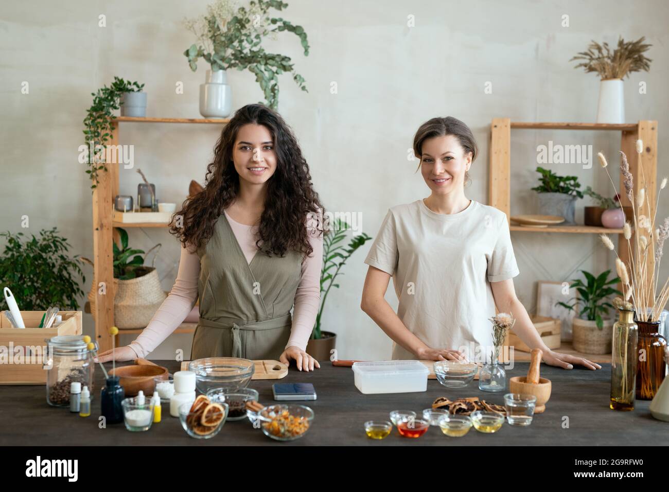 Two young females standing by table while one of them cutting hard soap mass in plastic container before making natural cosmetic products Stock Photo