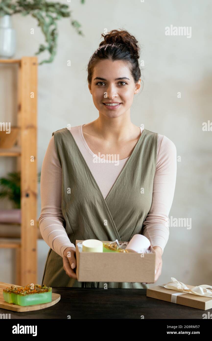 Young cheerful female in smart casualwear holding cardboard giftbox with handmade packed present while standing by table in front of camera Stock Photo