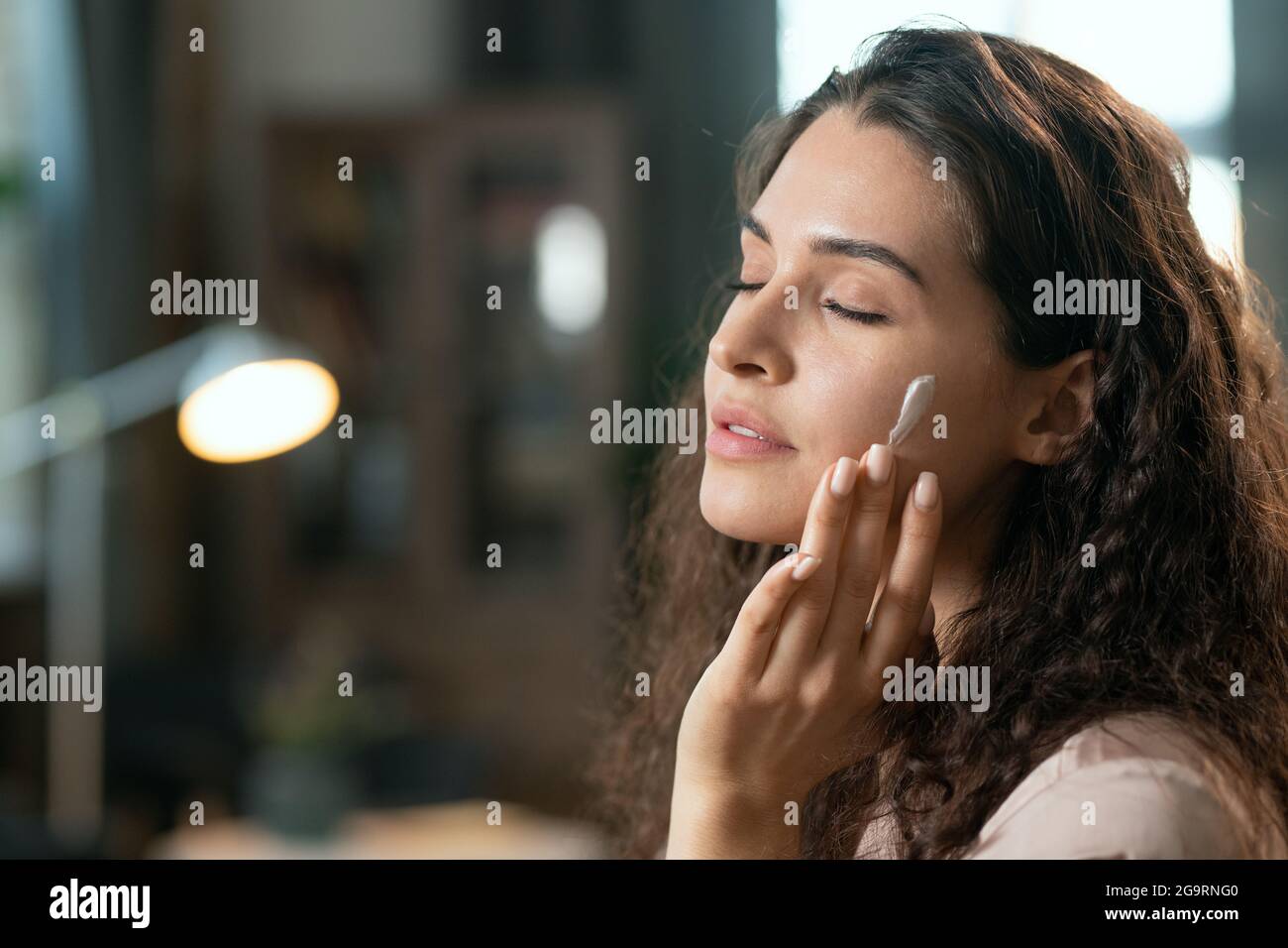 Young brunette pleased woman in silk pajamas applying natural cosmetic product on her face and enjoying it in the evening before sleep Stock Photo