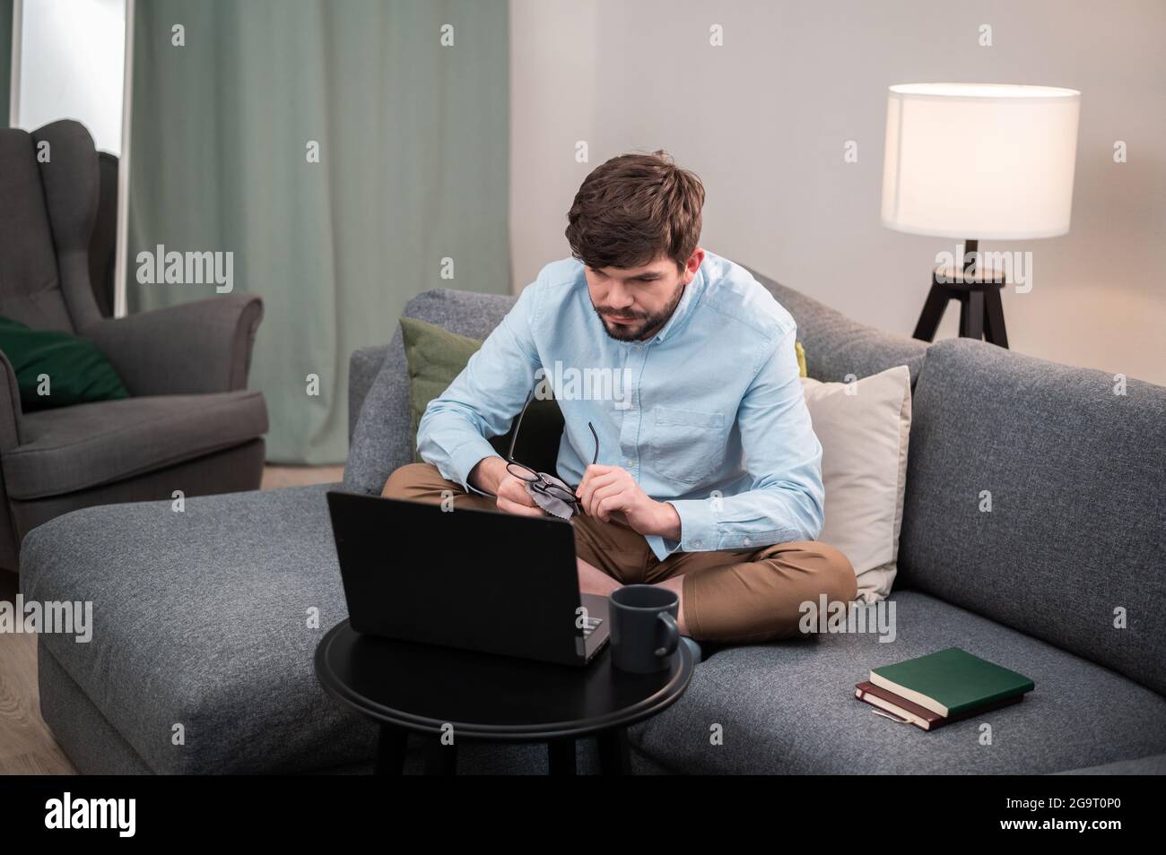 Hard work with the help of a young man's laptop, he sits on the couch, makes a report and drinks an invigorating coffee. Stock Photo