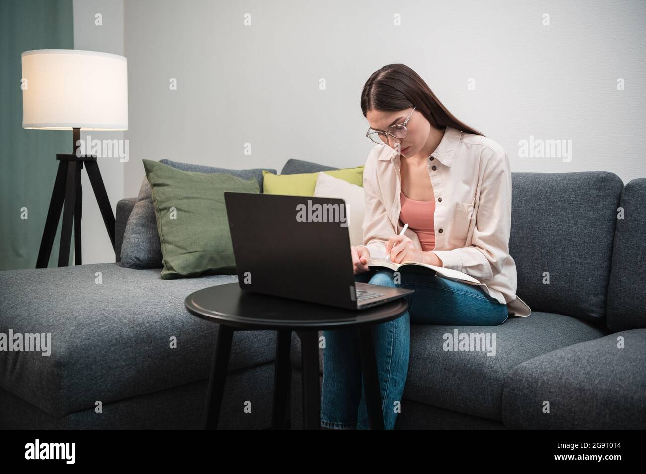 A young specialist girl is at a remote job, she is sitting at home on the couch, on the table next to her is a laptop. Stock Photo