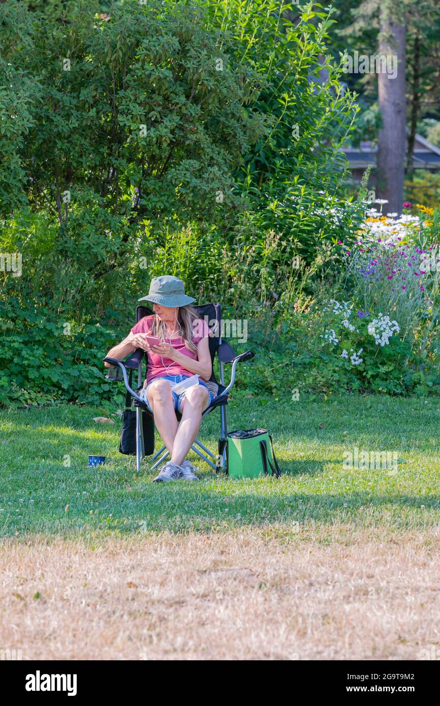 elderly woman with a hat sitting on a folding chair in the park holding smartphone and listening music. White Rock, BC, Canada-July 2,2021. Street vie Stock Photo