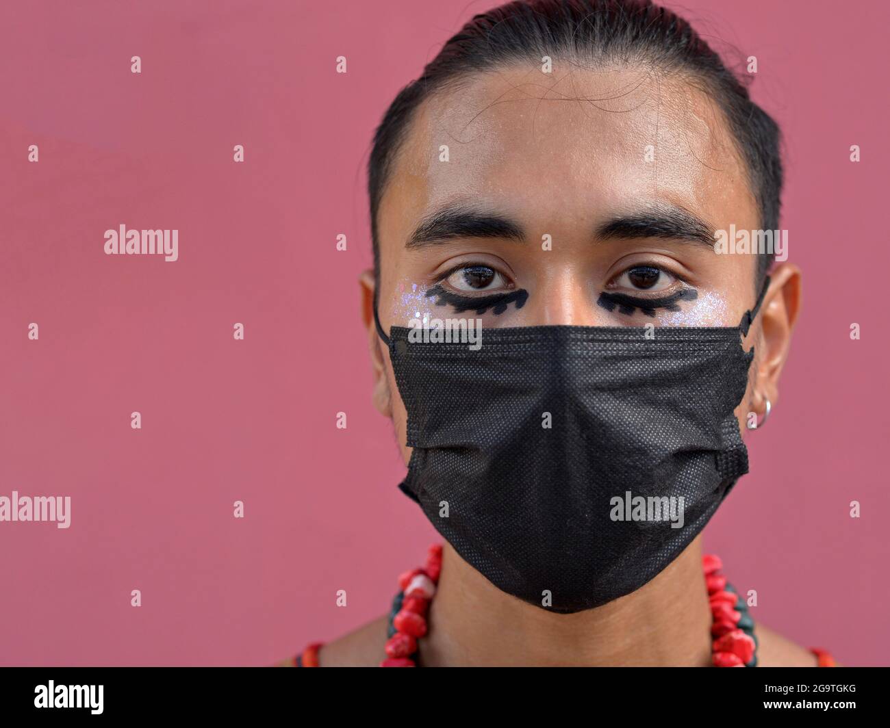 Young Latino man with romantic brown eyes and elaborate black eye make-up wears a disposable black corona face mask and looks at the viewer. Stock Photo