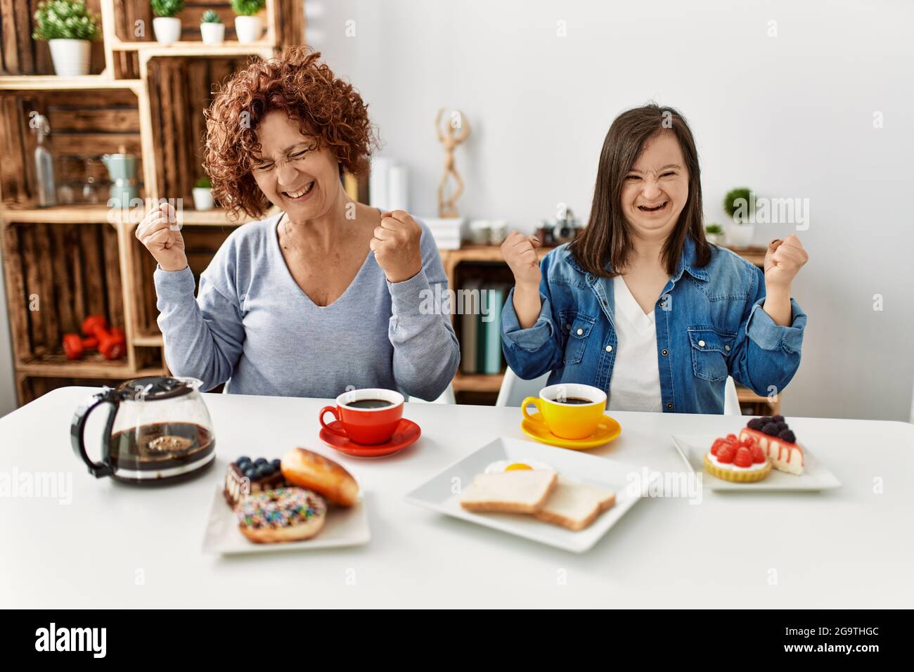 Family of mother and down syndrome daughter sitting at home eating breakfast very happy and excited doing winner gesture with arms raised, smiling and Stock Photo