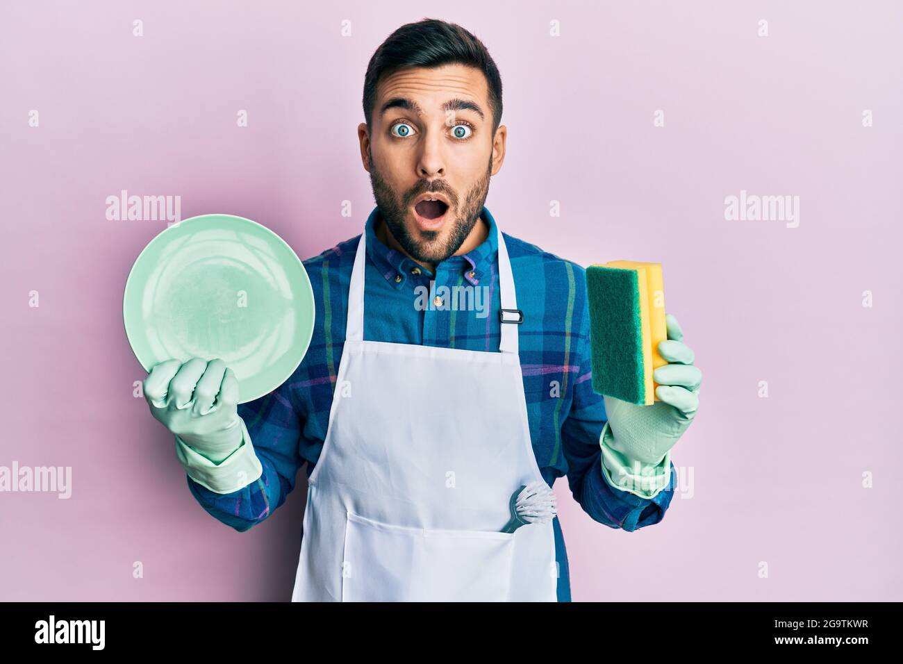 Young hispanic man wearing apron holding scourer washing dishes afraid