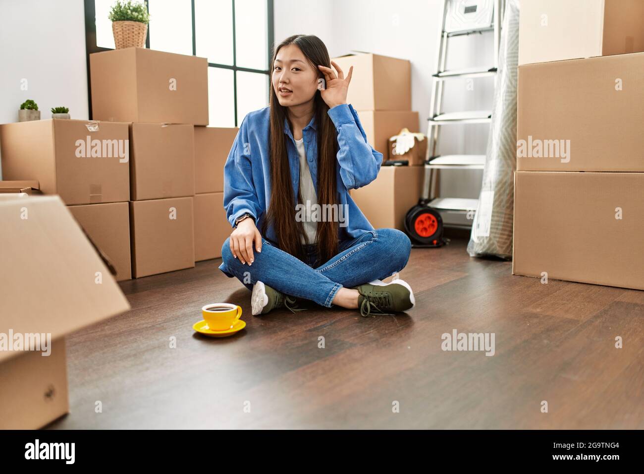 Young chinese girl sitting on the floor at new home smiling with hand ...