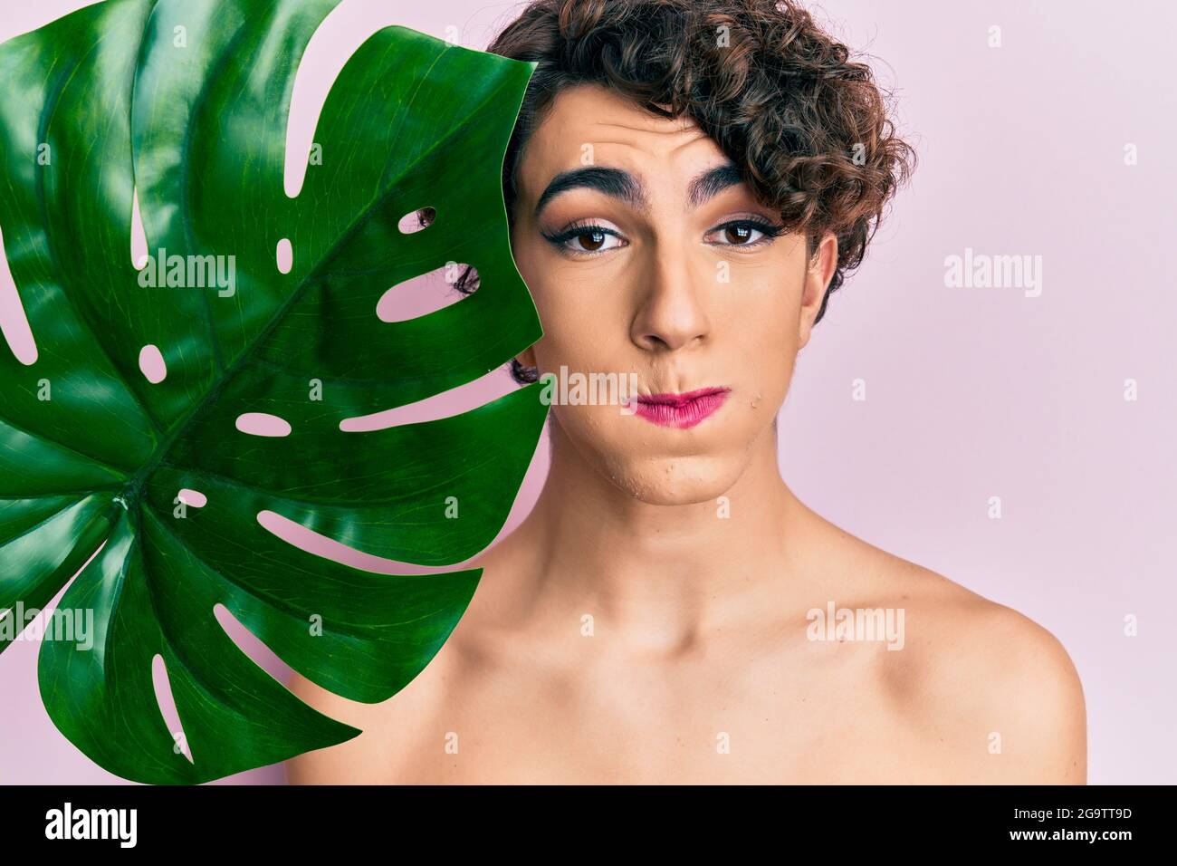 Young man wearing woman make up holding green plant leaf close to beautiful face puffing cheeks with funny face. mouth inflated with air, catching air Stock Photo