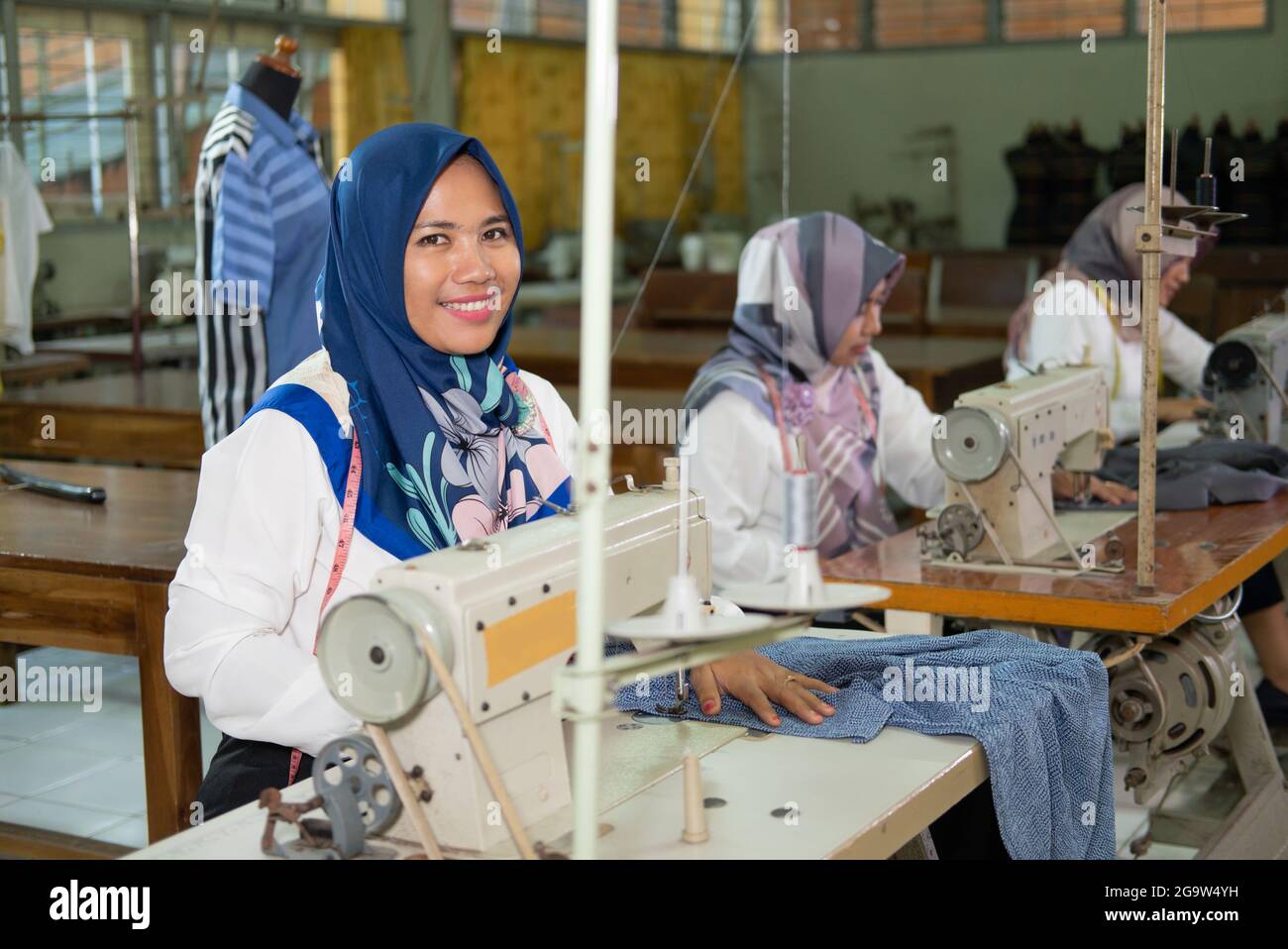 female employees in headscarves smile looking at cameras while working on sewing machines Stock Photo