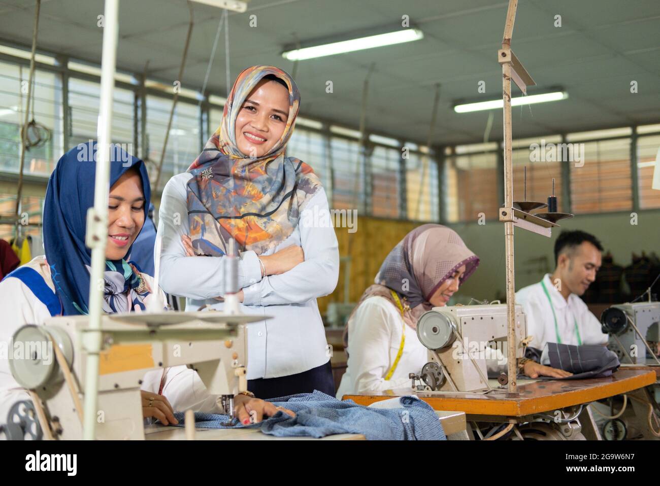female supervisor in the headscarf smiling crossed hands stands to the side the seamstress using the sewing machine Stock Photo