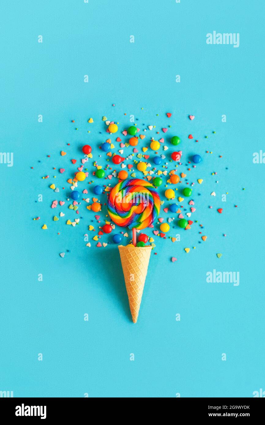 Ice cream waffle cone with colorful lollipop on stick, scattering of multicolored sweets and confectionery topping on blue background. Greeting card, Stock Photo