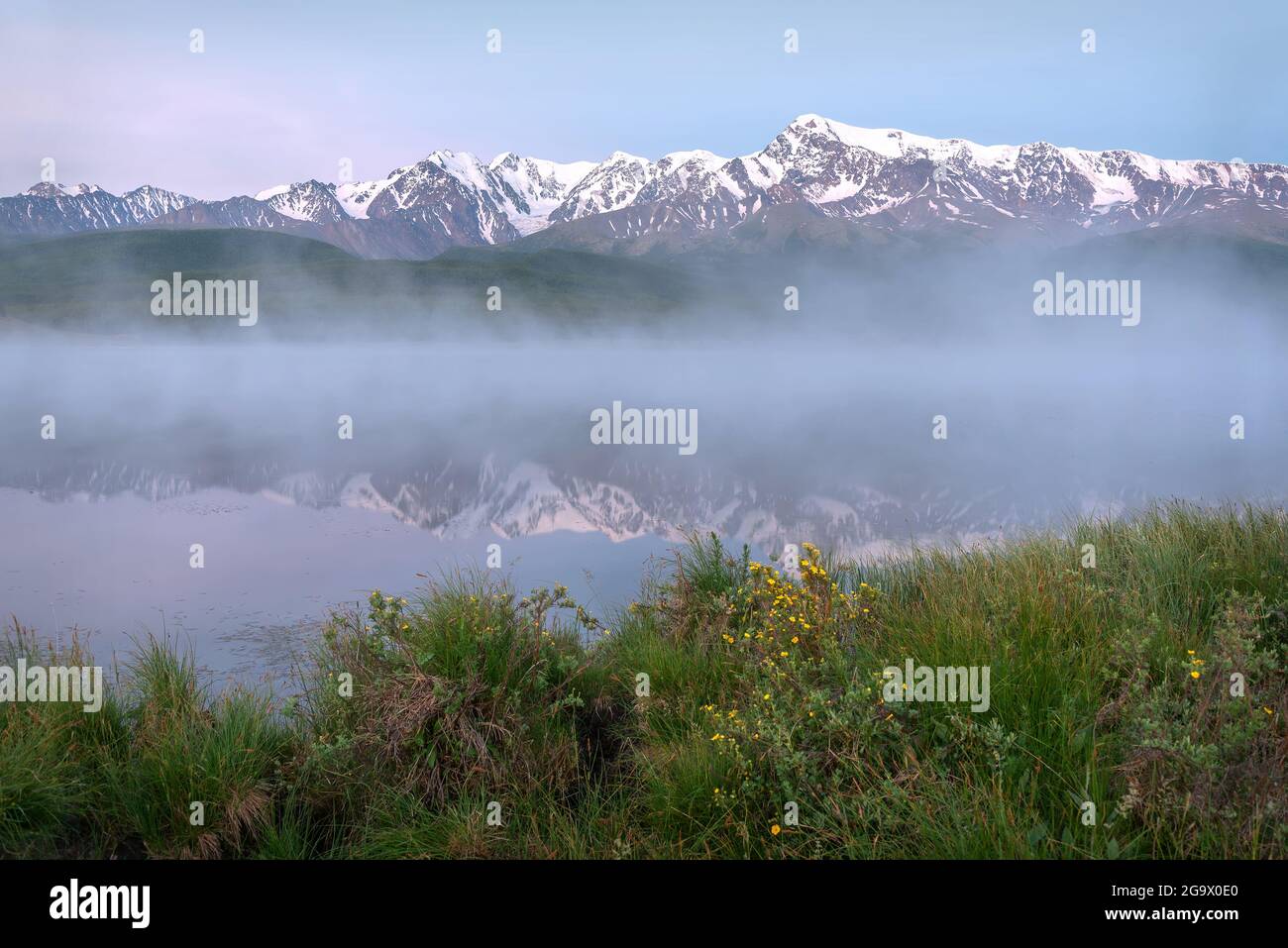Amazing delicate sunrise over the mountains covered with snow and forest, fog over the lake and beautiful reflections in the water against the blue sk Stock Photo