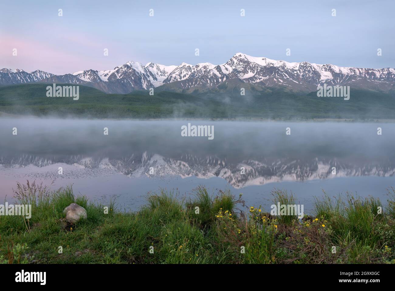 Amazing delicate sunrise over the mountains covered with snow and forest, fog over the lake and beautiful reflections in the water against the blue sk Stock Photo