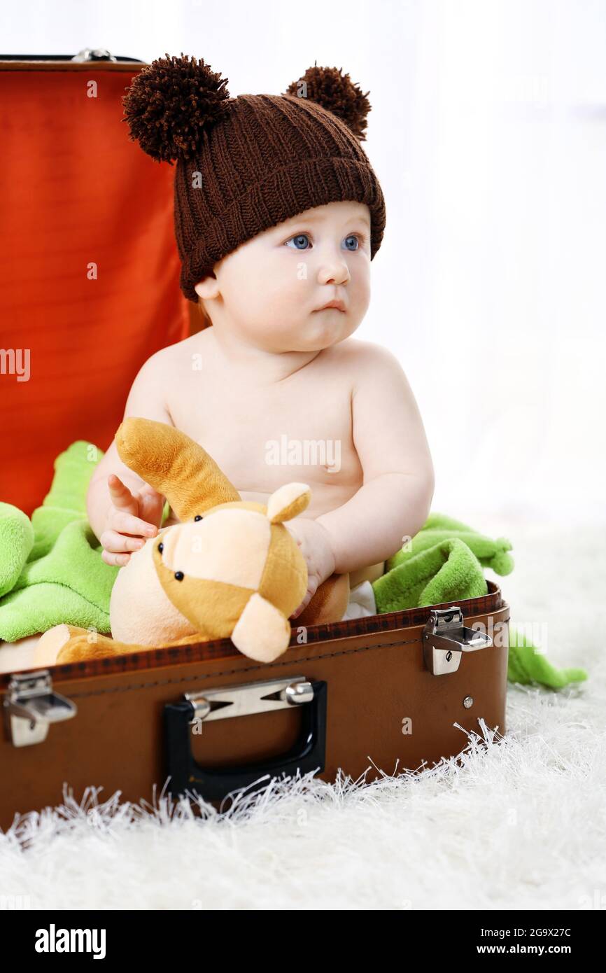 Cute baby boy in funny cap sitting in big suitcase on carpet, on light background Stock Photo