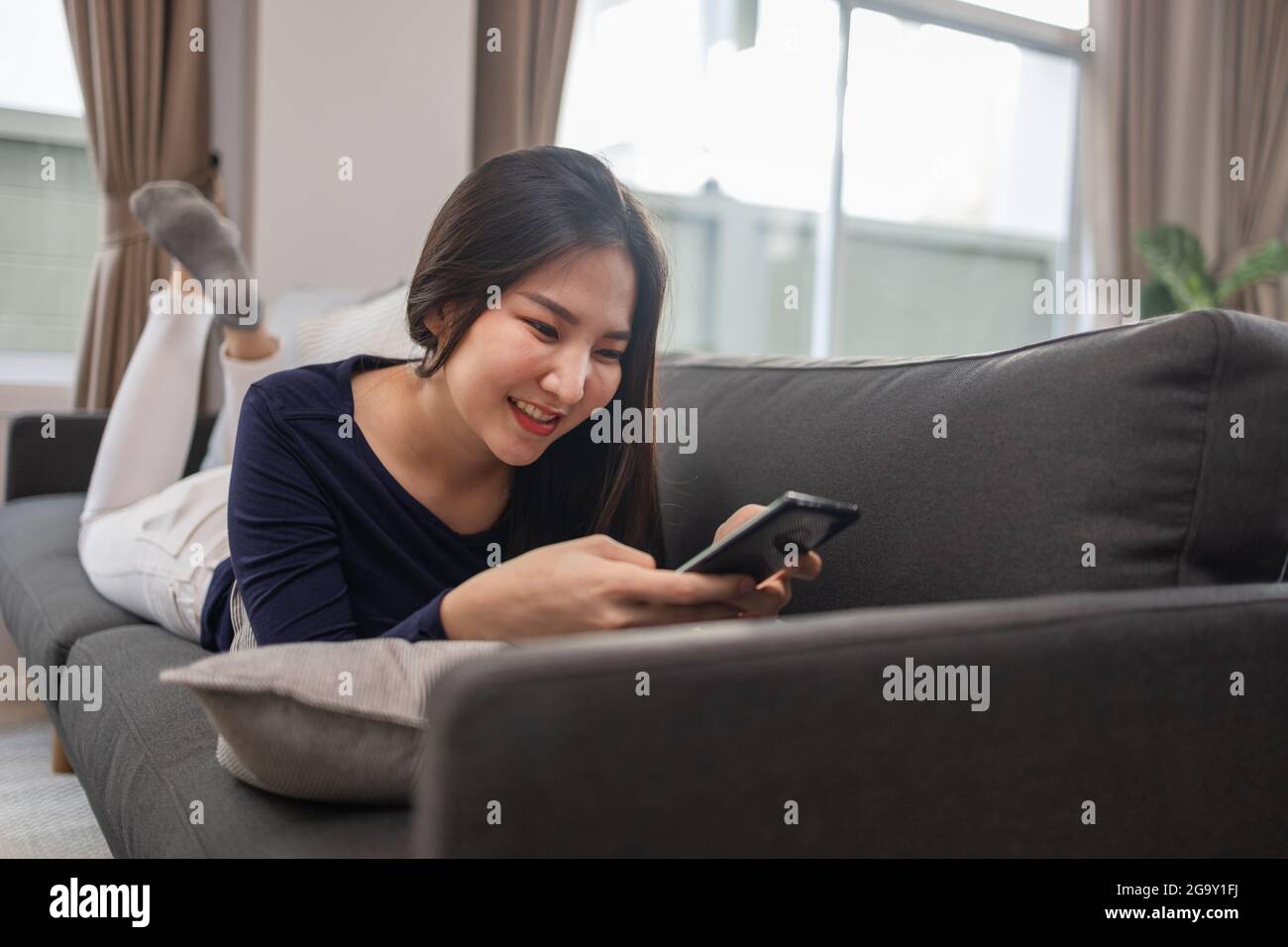 Work from home concept a young woman laying on a gray sofa in a living room surfing the internet. Stock Photo
